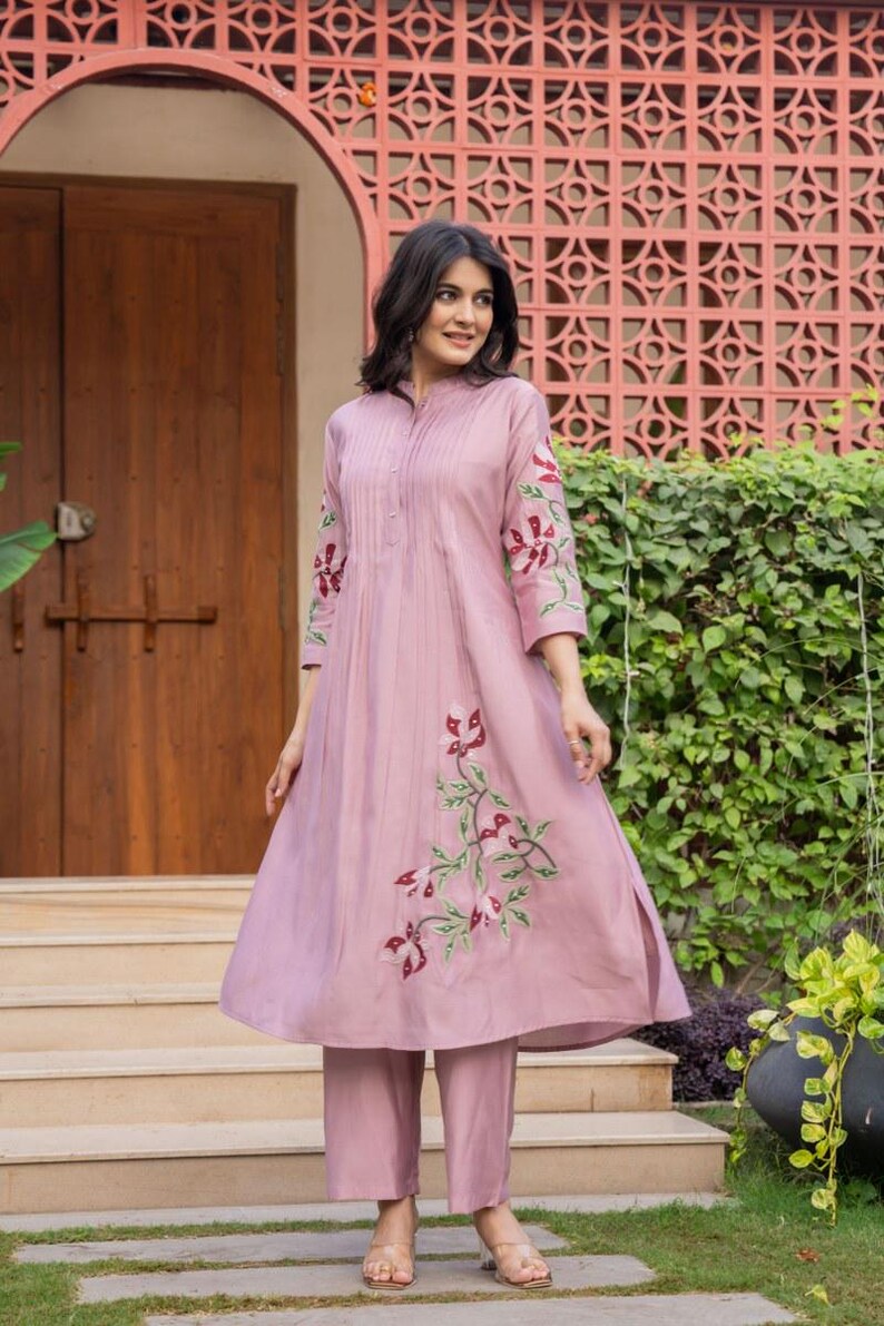 May include: A mauve-colored A-line kurta with three-quarter sleeves and matching straight-leg trousers. The garment features vertical pleats along the chest and embroidered floral motifs in shades of red, white, and green on the sleeves and skirt.