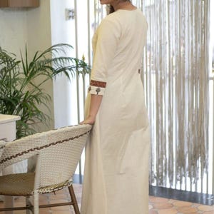 May include: A long, off-white, straight-cut tunic featuring three-quarter length sleeves with dark brown and black patterned cuffs. The tunic is paired with matching wide-leg trousers that have a dark brown and black geometric print at the hem.