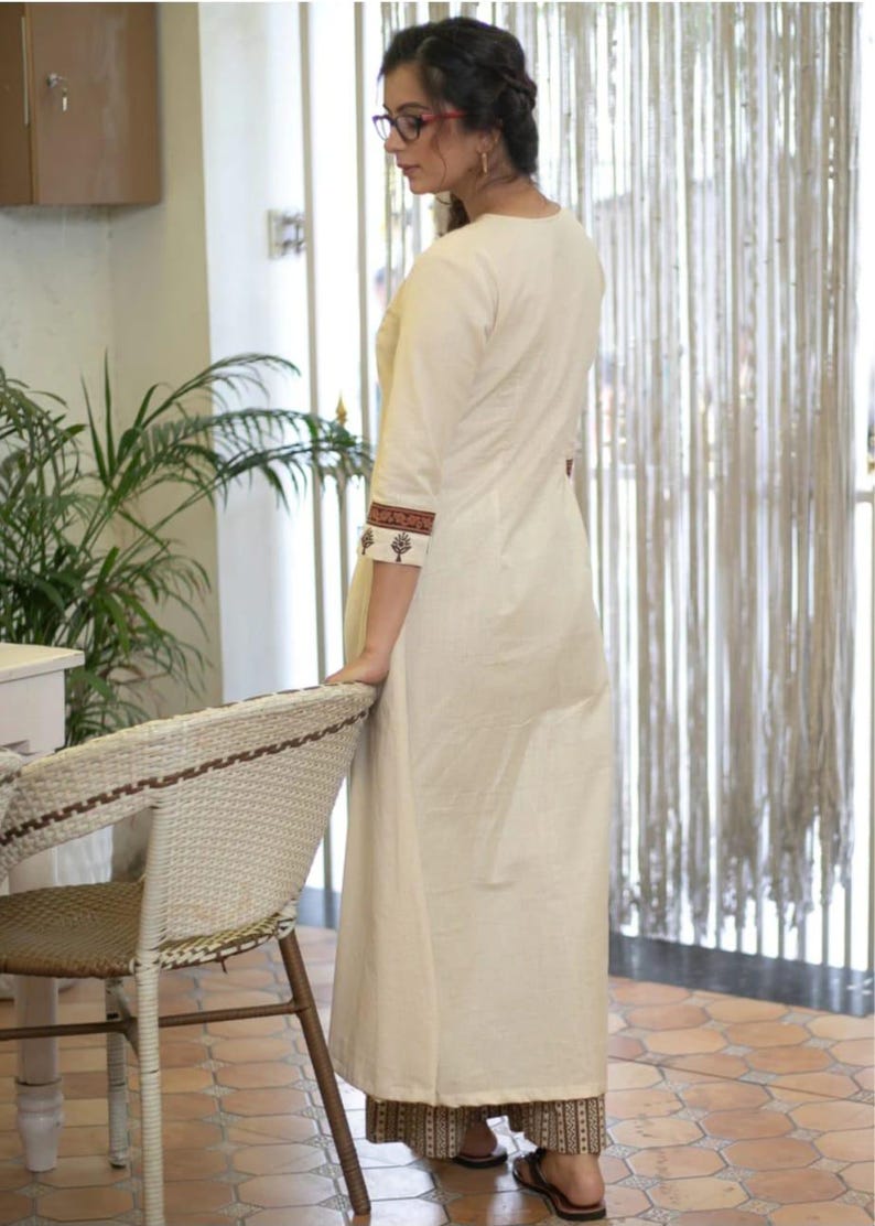 May include: A long, off-white, straight-cut tunic featuring three-quarter length sleeves with dark brown and black patterned cuffs. The tunic is paired with matching wide-leg trousers that have a dark brown and black geometric print at the hem.