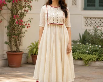 May include: A cream-colored A-line dress featuring a square chest panel adorned with small circular mirrors and red trim, paired with solid dark red wide-leg trousers. The dress has three-quarter length sleeves and a round neckline.
