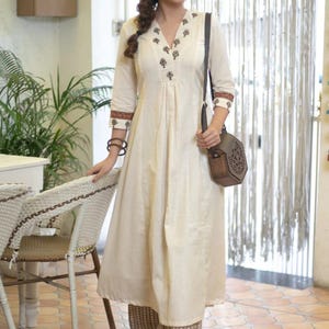 May include: An off-white A-line kurta dress featuring a V-neckline with dark brown floral embroidery, three-quarter sleeves with patterned cuffs, and matching wide-leg trousers. A dark brown hexagonal wooden crossbody bag is worn over the shoulder.