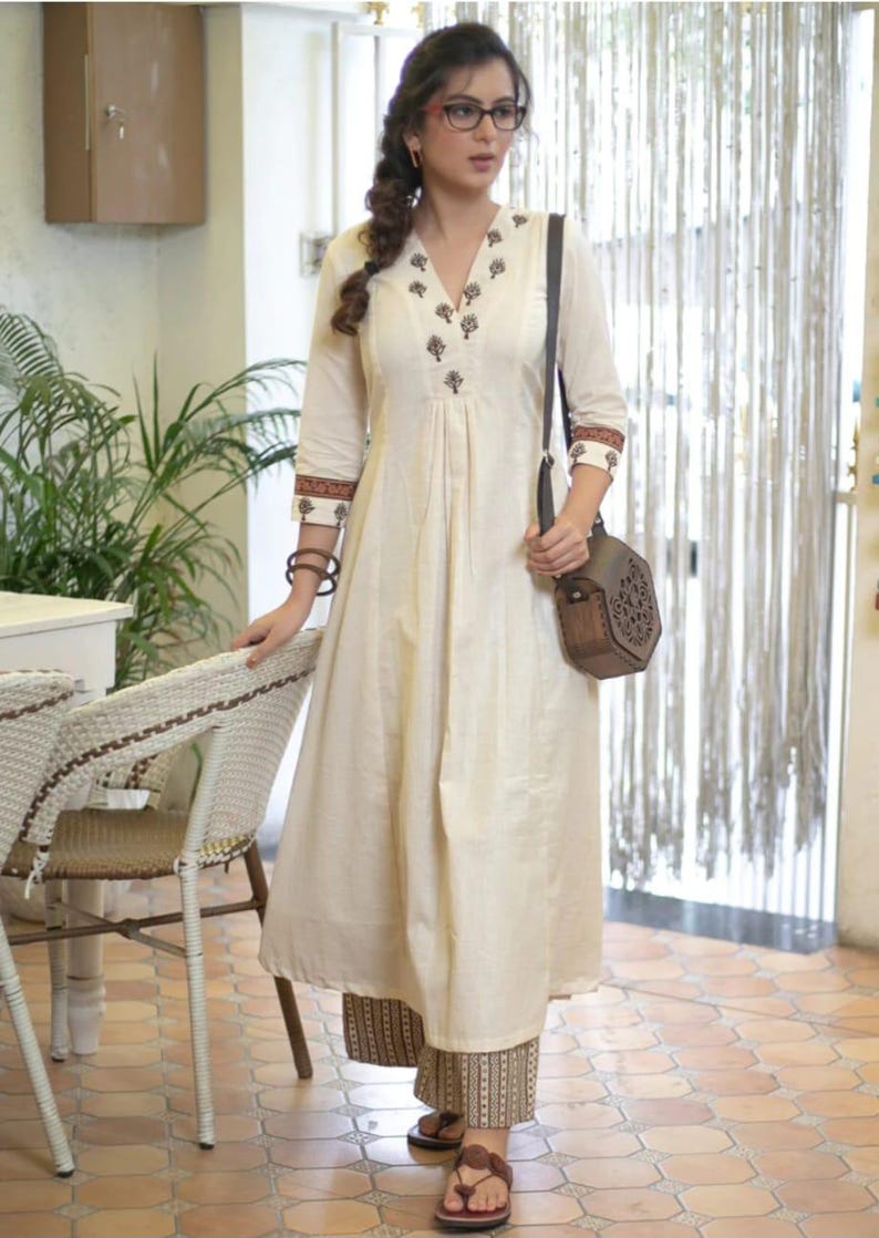 May include: An off-white A-line kurta dress featuring a V-neckline with dark brown floral embroidery, three-quarter sleeves with patterned cuffs, and matching wide-leg trousers. A dark brown hexagonal wooden crossbody bag is worn over the shoulder.