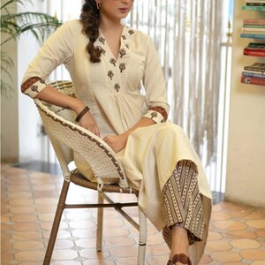 May include: A cream-coloured long-sleeved tunic featuring a V-neckline with dark brown floral embroidery, paired with brown and cream patterned wide-leg trousers. The tunic has matching brown floral trim on the cuffs and a pleated front bodice.