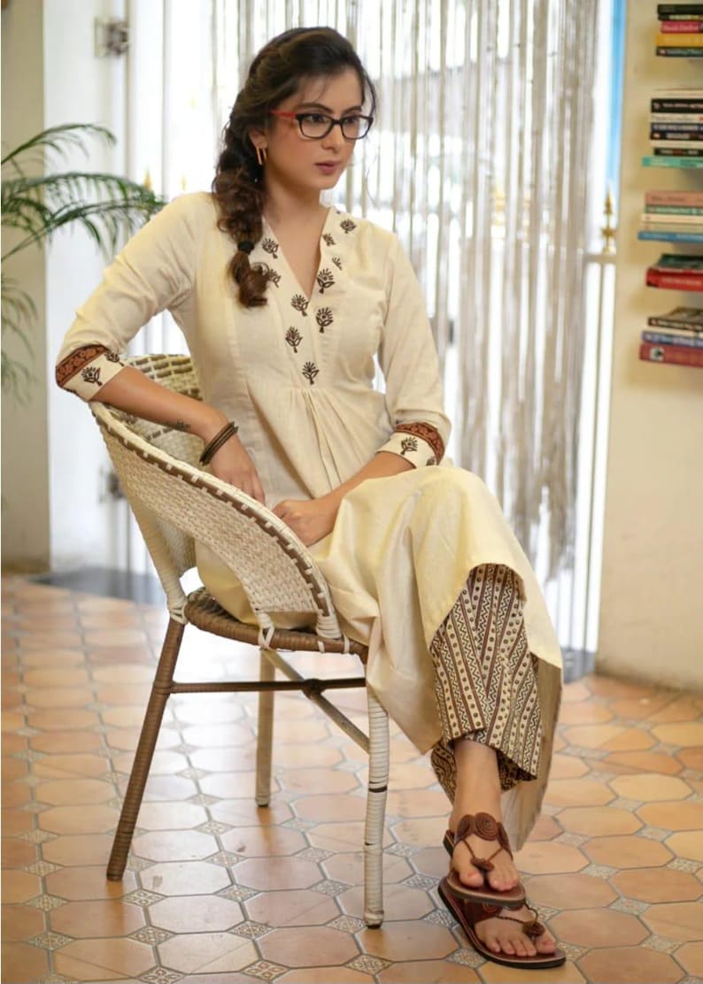 May include: A cream-coloured long-sleeved tunic featuring a V-neckline with dark brown floral embroidery, paired with brown and cream patterned wide-leg trousers. The tunic has matching brown floral trim on the cuffs and a pleated front bodice.