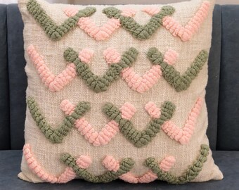Könnte beinhalten: Quadratisches Zierkissen mit cremefarbenem Leinenstoff. Das Kissen hat ein sich wiederholendes Muster aus rosa und grünen strukturierten Chevron-Designs. Das Kissen liegt auf einem blauen Sofa.
