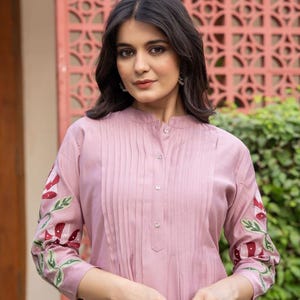 May include: A mauve-colored tunic featuring a mandarin collar, a button-down front placket with vertical pleats, and three-quarter length sleeves adorned with embroidered floral patterns in shades of red, white, and green on the outer arms.