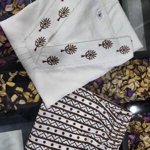 May include: A white tunic featuring a V-neckline with brown floral embroidery and a matching pair of white trousers with a brown geometric pattern of horizontal lines and dots. The garment includes a small white tag with the number 40 printed on it.
