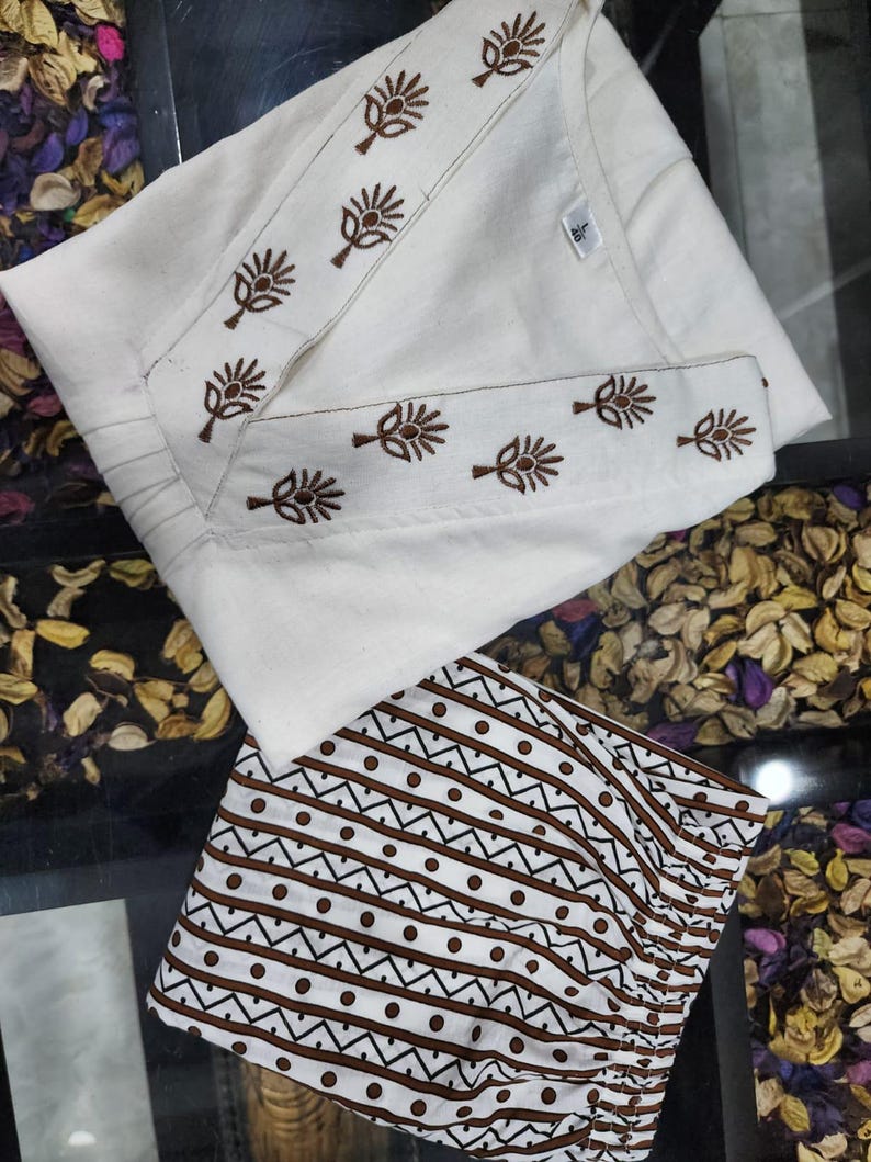 May include: A white tunic featuring a V-neckline with brown floral embroidery and a matching pair of white trousers with a brown geometric pattern of horizontal lines and dots. The garment includes a small white tag with the number 40 printed on it.