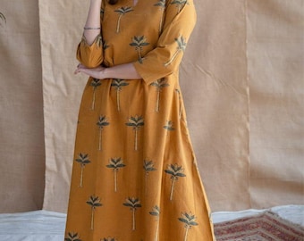 Könnte beinhalten: Ein senfgelbes, wadenlanges Kleid mit einem sich wiederholenden, dunkelgrünen Palmenmuster, Dreiviertelärmeln und einem V-Ausschnitt. Das Kleid hat einen ausgestellten Rock, wird über einem weißen Unterrock getragen und mit metallischen Sandalen kombiniert.