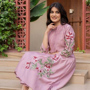 May include: A mauve midi dress featuring a mandarin collar, vertical pleats on the bodice, and three-quarter length sleeves. The dress is adorned with embroidered floral motifs in shades of red, white, and green on the sleeves and skirt.