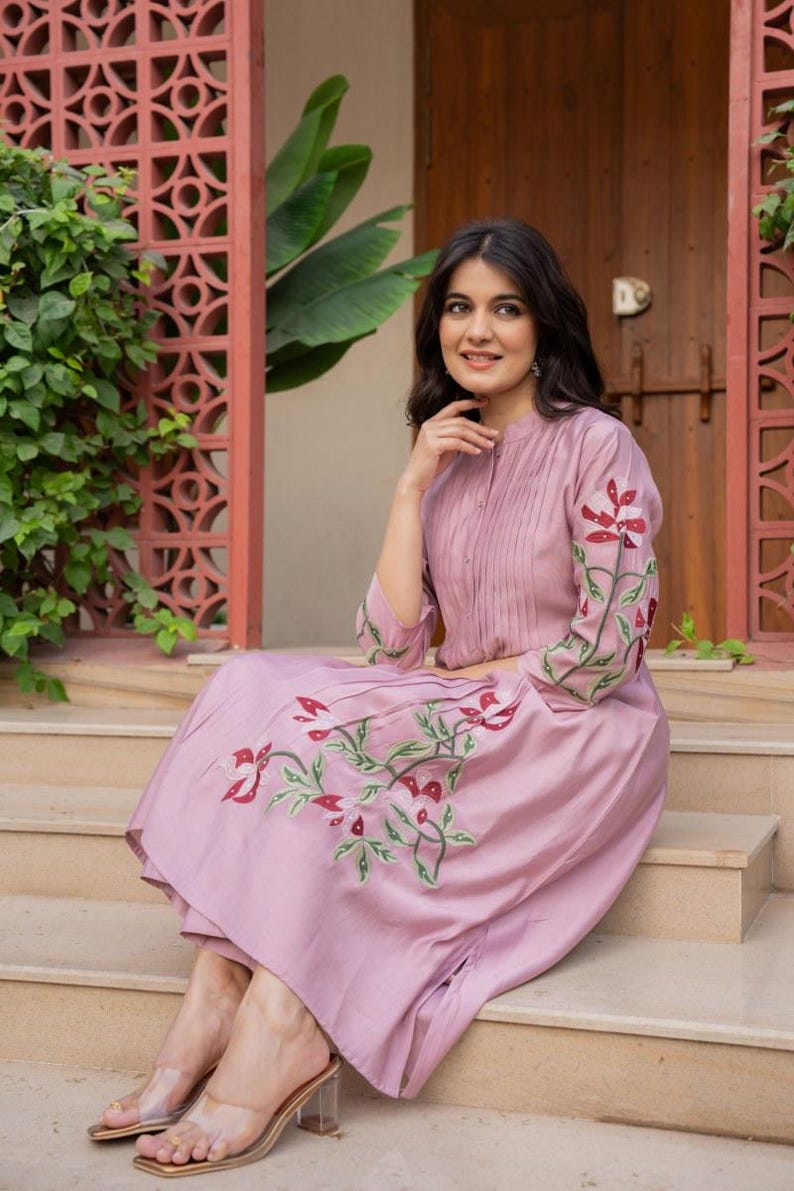 May include: A mauve midi dress featuring a mandarin collar, vertical pleats on the bodice, and three-quarter length sleeves. The dress is adorned with embroidered floral motifs in shades of red, white, and green on the sleeves and skirt.