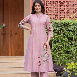 May include: A mauve knee-length tunic with a mandarin collar, vertical pleats on the bodice, and three-quarter sleeves. The garment features floral embroidery in shades of red, white, and green on the sleeves and lower side, paired with matching trousers.