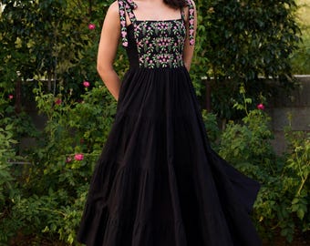 Könnte beinhalten: Ein schwarzes Midikleid mit quadratischem Ausschnitt und einem Oberteil, das mit kleinen rosa Blumen und grünen Blättern bestickt ist. Das Kleid hat breite Schulterträger zum Binden und einen gestuften, ausgestellten Rock, der unter die Knie reicht.