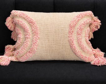 Könnte beinhalten: Ein rechteckiges beiges Kissen mit rosa Akzenten. Das Kissen hat einen strukturierten Stoff mit rosa, halbkreisförmigen Designs auf jeder Seite. Rosa Quasten schmücken die Enden. Ideal als dekoratives Element für ein Sofa oder Bett. Ein stilvolles Wohnaccessoire.