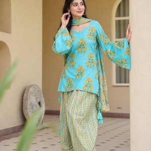 Könnte beinhalten: Eine leuchtend türkisfarbene Tunika mit gelbem Blumenmuster, V-Ausschnitt und langen, ausgestellten Ärmeln. Die passende Hose im Dhoti-Stil zeigt ein vertikales gelb-weißes Zickzackmuster. Ein dazu passender türkisfarbener Schal mit gelben Blumenakzenten vervollständigt das Set.