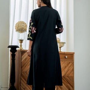 May include: A long, black, knee-length tunic featuring three-quarter length sleeves with embroidered floral patterns in shades of pink, burgundy, and green. The garment is paired with matching black trousers and has a simple, straight-cut silhouette.