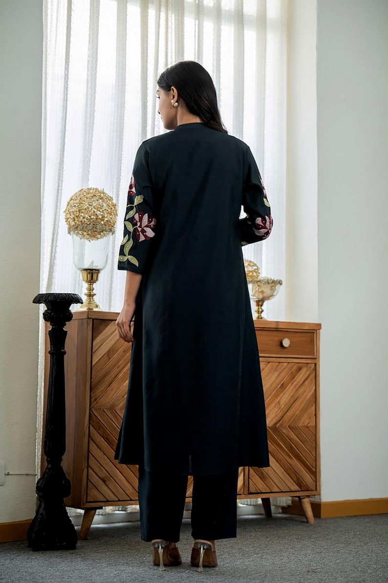 May include: A long, black, knee-length tunic featuring three-quarter length sleeves with embroidered floral patterns in shades of pink, burgundy, and green. The garment is paired with matching black trousers and has a simple, straight-cut silhouette.