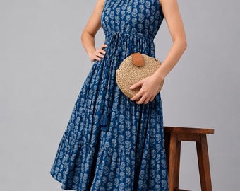 Könnte beinhalten: Ein ärmelloses Midikleid in Dunkelblau mit weißem floralem Blockdruckmuster, rundem Ausschnitt, einer Taille mit Kordelzug und Quastendetails sowie einem gestuften Rock, kombiniert mit einer runden Handtasche aus geflochtenem Stroh.