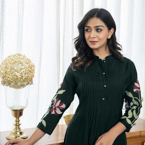 May include: A dark green long-sleeved dress featuring a mandarin collar, a button-down front placket with vertical pleats, and floral embroidery in shades of pink, white, and green on the sleeves and lower portion of the garment.