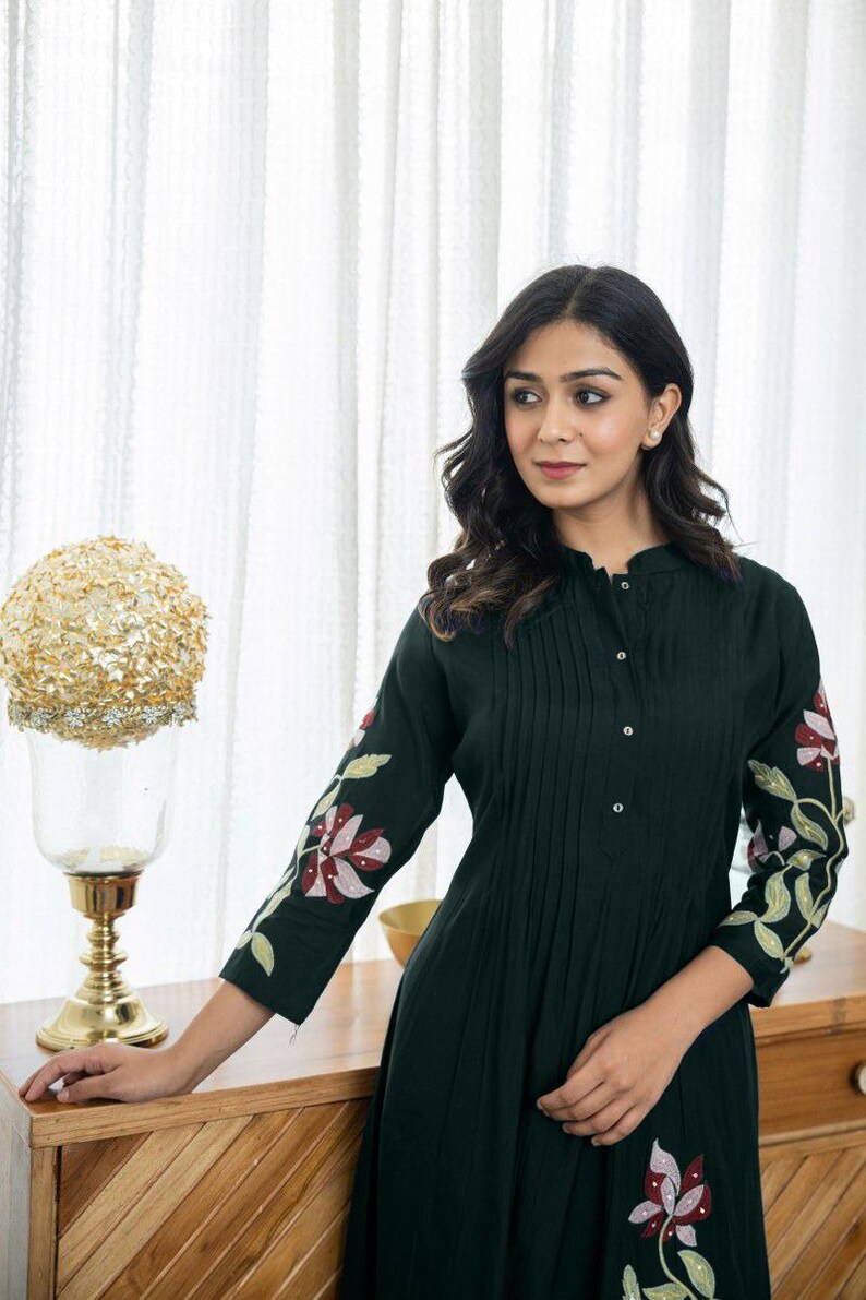 May include: A dark green long-sleeved dress featuring a mandarin collar, a button-down front placket with vertical pleats, and floral embroidery in shades of pink, white, and green on the sleeves and lower portion of the garment.