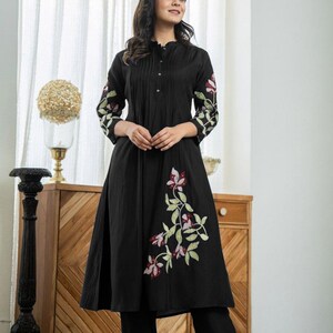 May include: A black knee-length tunic with a mandarin collar and button-down front, featuring three-quarter sleeves and a flared skirt. The garment is decorated with embroidered floral patterns in shades of pink, white, and green on the sleeves and side.