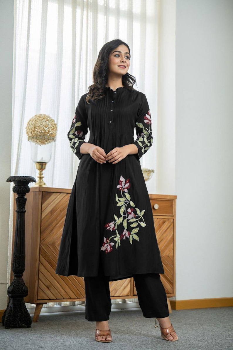 May include: A black knee-length tunic with a mandarin collar and button-down front, featuring three-quarter sleeves and a flared skirt. The garment is decorated with embroidered floral patterns in shades of pink, white, and green on the sleeves and side.