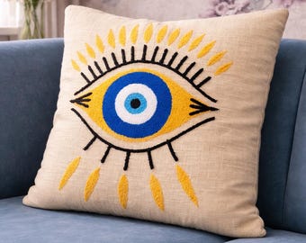 Könnte beinhalten: Beiges quadratisches Zierkissen mit einem gestickten bösen Blick-Design. Das Auge hat eine blaue Iris, gelbe und schwarze Details, vor einem hellblauen Sofa. Ein Einrichtungsgegenstand.