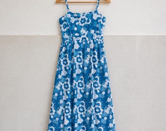Könnte beinhalten: Ein ärmelloses Midikleid mit einem blauen und weißen Blumenmuster. Das Kleid hat dünne Träger, einen eckigen Ausschnitt und ein körpernah geschnittenes Oberteil, das in einen fließenden Rock übergeht, präsentiert auf einem Holzkleiderbügel.