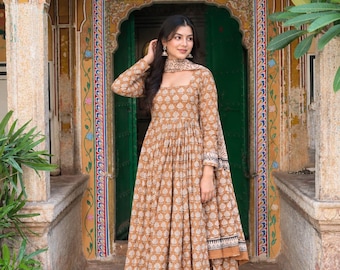 Könnte beinhalten: Ein beigefarbener Anarkali-Anzug mit einem sich wiederholenden weißen Blumenmuster im Blockdruck. Das langärmelige Kleid hat einen eckigen Ausschnitt, eine geraffte Taille und einen metallisch goldenen Saum sowie eine passende Hose und Dupatta.