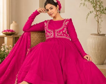 Könnte beinhalten: Ein leuchtend fuchsiafarbenes, bodenlanges Kleid mit langen Ärmeln, rundem Ausschnitt und einem eng anliegenden Oberteil, das mit aufwendigen Goldstickereien verziert ist. Das Kleid hat einen weiten, voluminösen Rock und einen passenden fuchsiafarbenen Dupatta mit Goldrand.
