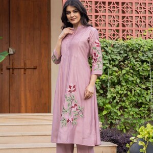 May include: A mauve-colored kurta-style tunic featuring vertical pintuck pleats at the chest, three-quarter length sleeves, and floral embroidery in shades of red and green. The tunic is paired with matching mauve straight-leg trousers.