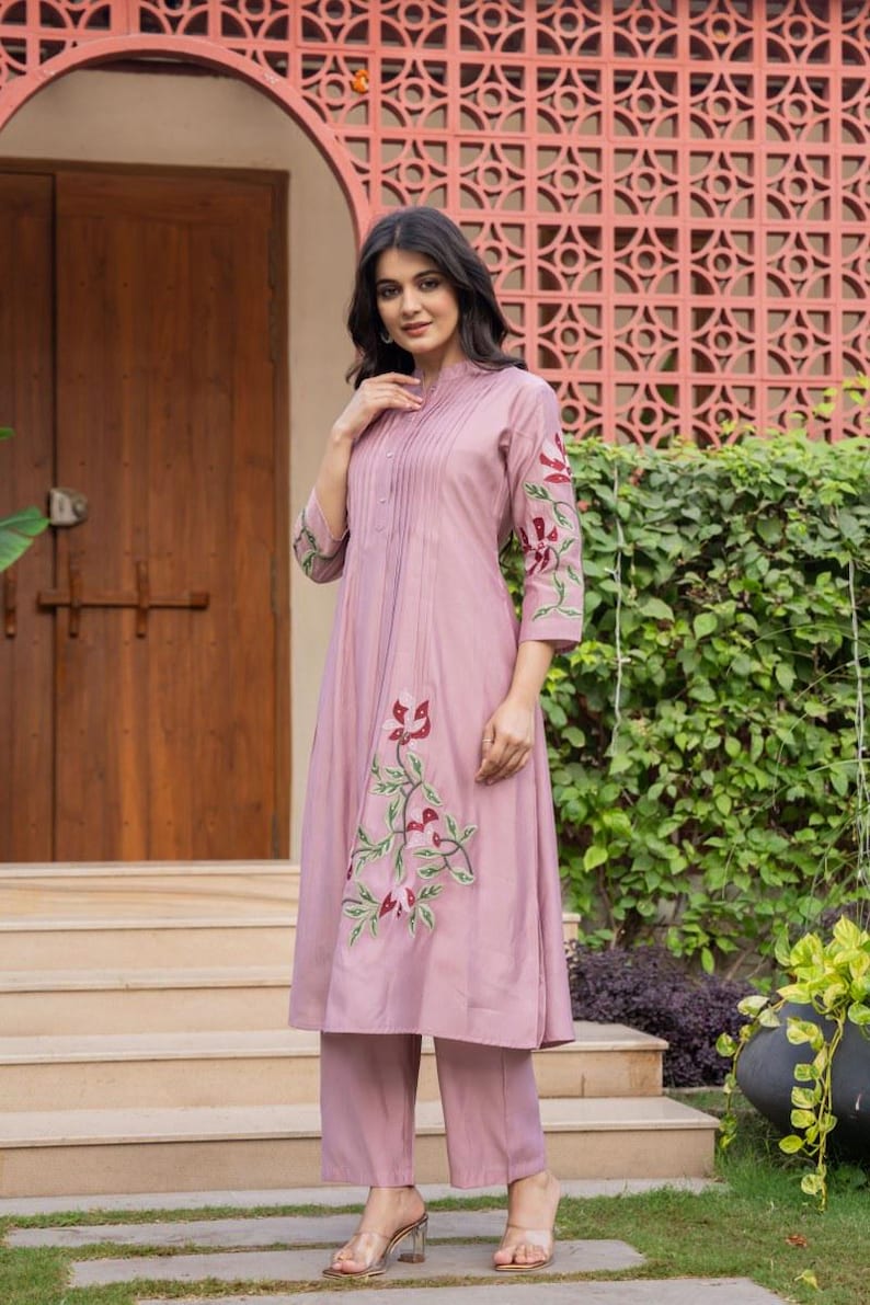 May include: A mauve-colored kurta-style tunic featuring vertical pintuck pleats at the chest, three-quarter length sleeves, and floral embroidery in shades of red and green. The tunic is paired with matching mauve straight-leg trousers.