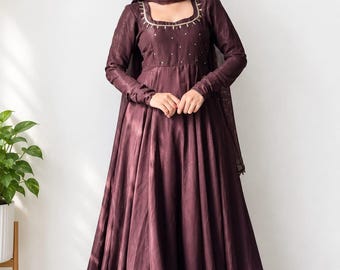 Könnte beinhalten: Ein bodenlanges, langärmeliges Kleid in Dunkelpflaume mit quadratischem Ausschnitt, der mit goldfarbenen Stickereien und kleinen Verzierungen versehen ist. Das Kleid hat einen weiten, plissierten Rock und einen passenden, transparenten Schal.