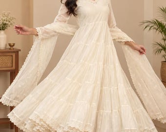 Könnte beinhalten: Ein cremefarbenes Anarkali-Kleid mit V-Ausschnitt, langen, transparenten Ärmeln und einem mehrstufigen, ausgestellten Rock. Das Kleidungsstück ist mit feiner Spitzenborte an Saum und Ärmeln verziert und wird mit einer passenden Hose und Dupatta kombiniert.