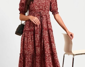 Könnte beinhalten: Ein wadenlanges Kleid in tiefem Kastanienbraun mit einem sich wiederholenden cremefarbenen Blumen- und Paisleymuster. Das Kleid hat einen V-Ausschnitt, kurze Puffärmel mit elastischen Bündchen, eine betonte Taille mit Bindeband und einen Stufenrock.