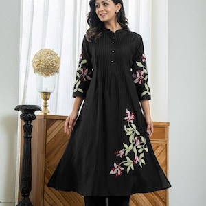 May include: A black knee-length tunic dress featuring a mandarin collar, a button-down front placket with vertical pleats, and three-quarter length sleeves. The garment is decorated with embroidered floral motifs in shades of pink, red, and green.