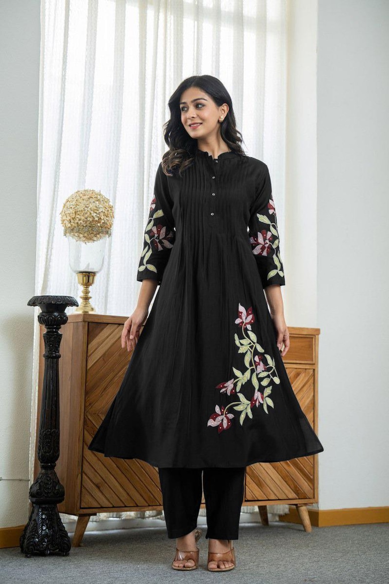 May include: A black knee-length tunic dress featuring a mandarin collar, a button-down front placket with vertical pleats, and three-quarter length sleeves. The garment is decorated with embroidered floral motifs in shades of pink, red, and green.