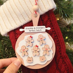May include: A wooden Christmas ornament featuring gingerbread people and the text "Grandma & Grandpa's Perfect Batch." The ornament is hanging on a red stocking. The gingerbread people have names on them.