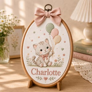 May include: An oval-shaped, framed embroidery featuring a gray tabby kitten holding balloons. The kitten wears a pink bow and the name "Charlotte" is stitched below. The frame is wood-toned, with a pink bow at the top.