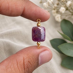 May include: A gold-colored jewelry component featuring a rectangular, deep purple gemstone. The gemstone is suspended between two gold beads and a small gold loop. The jewelry component is held between two fingers.