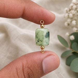 May include: A rectangular green gemstone bead with gold-colored accents. The bead is held between two fingers. The gemstone has dark green and light green marbling. The gold accents are small round beads and a loop at each end.