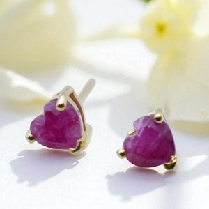 May include: A pair of heart-shaped ruby earrings with gold settings. The earrings are a vibrant pinkish-red color and are set against a white background with soft floral accents.