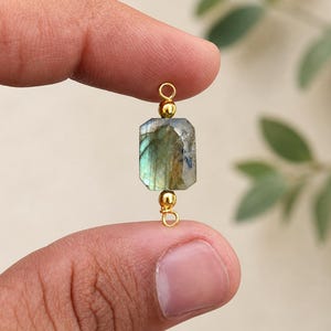 Può includere: Un pendente rettangolare in pietra di labradorite con dettagli color oro. La pietra preziosa presenta una miscela di tonalità verdi, blu e grigie. Il pendente è tenuto tra due dita.