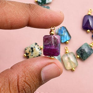 May include: A rectangular, multi-colored gemstone bead with gold-colored findings is held between fingers. Other gemstone beads in various colors, including purple, blue, and green, are visible in the background. The beads are likely for jewelry making.