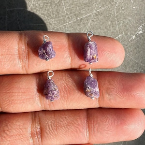 May include: Four raw, uncut purple gemstone pendants with silver wire loops for jewellery making. The gemstones have a rough, natural texture and varying shapes. The pendants are displayed against a neutral background.