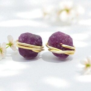 May include: A pair of raw, purple gemstone stud earrings. Each stone is wrapped with delicate gold-coloured wire. The earrings are set against a white background with small white flowers.