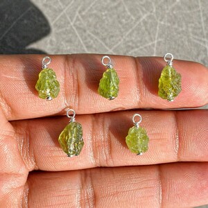 May include: Six raw peridot gemstone pendants. Each pendant features a rough, olive-green peridot stone with a silver-coloured wire loop for hanging. The pendants are displayed against a neutral background.