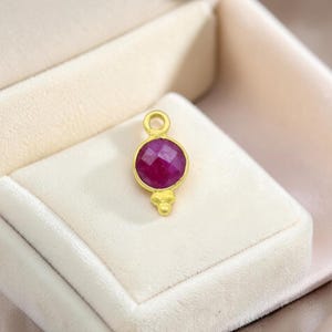 Könnte beinhalten: Ein goldfarbener Anhänger mit einem runden, facettierten, tiefroten Edelstein. Der Anhänger hat eine Schlaufe für eine Kette und ein dekoratives Element unten. Präsentiert auf einer weißen Samtoberfläche.