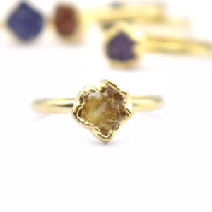 Könnte beinhalten: Eine Auswahl an goldenen Ringen mit verschiedenen Edelsteinen. Der Hauptring hat einen hellbraunen, unregelmäßig geformten Stein. Weitere Ringe im Hintergrund haben blaue, braune und violette Steine. Die Ringe sind vor weißem Hintergrund.