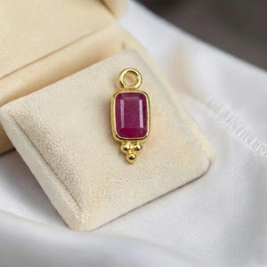 Puede incluir: Colgante de oro con una gema rectangular de color rojo intenso. El colgante tiene un pequeño lazo en la parte superior y tres pequeñas esferas doradas en la parte inferior. Presentado sobre un cojín crema dentro de un joyero.